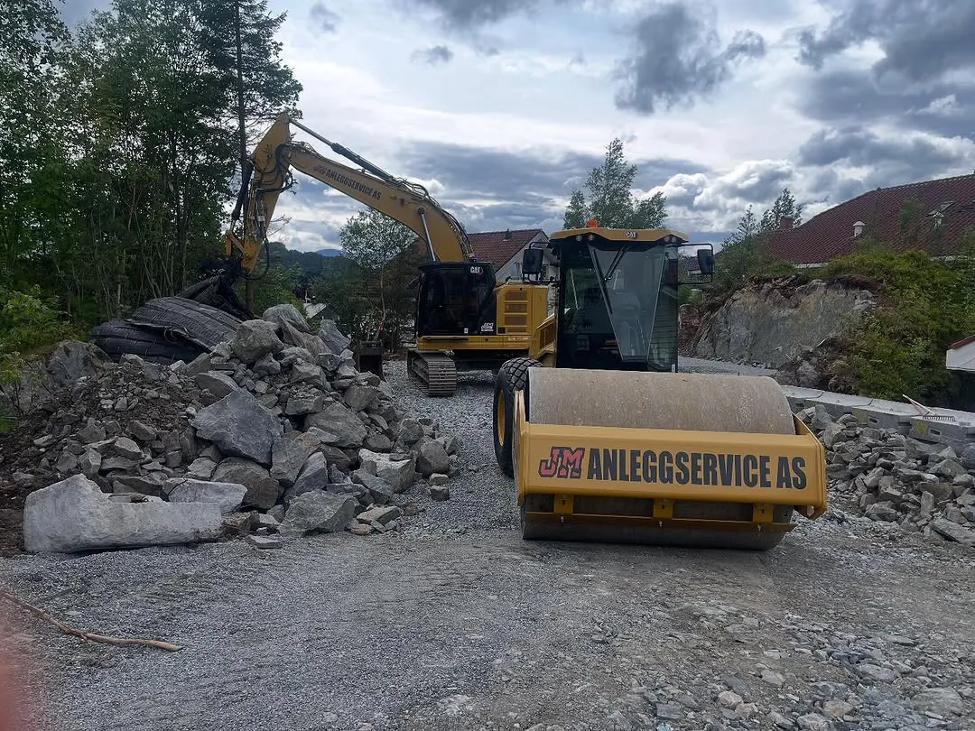 Vals med JM Anleggservice-logo på valsen, foran en gravemaskin